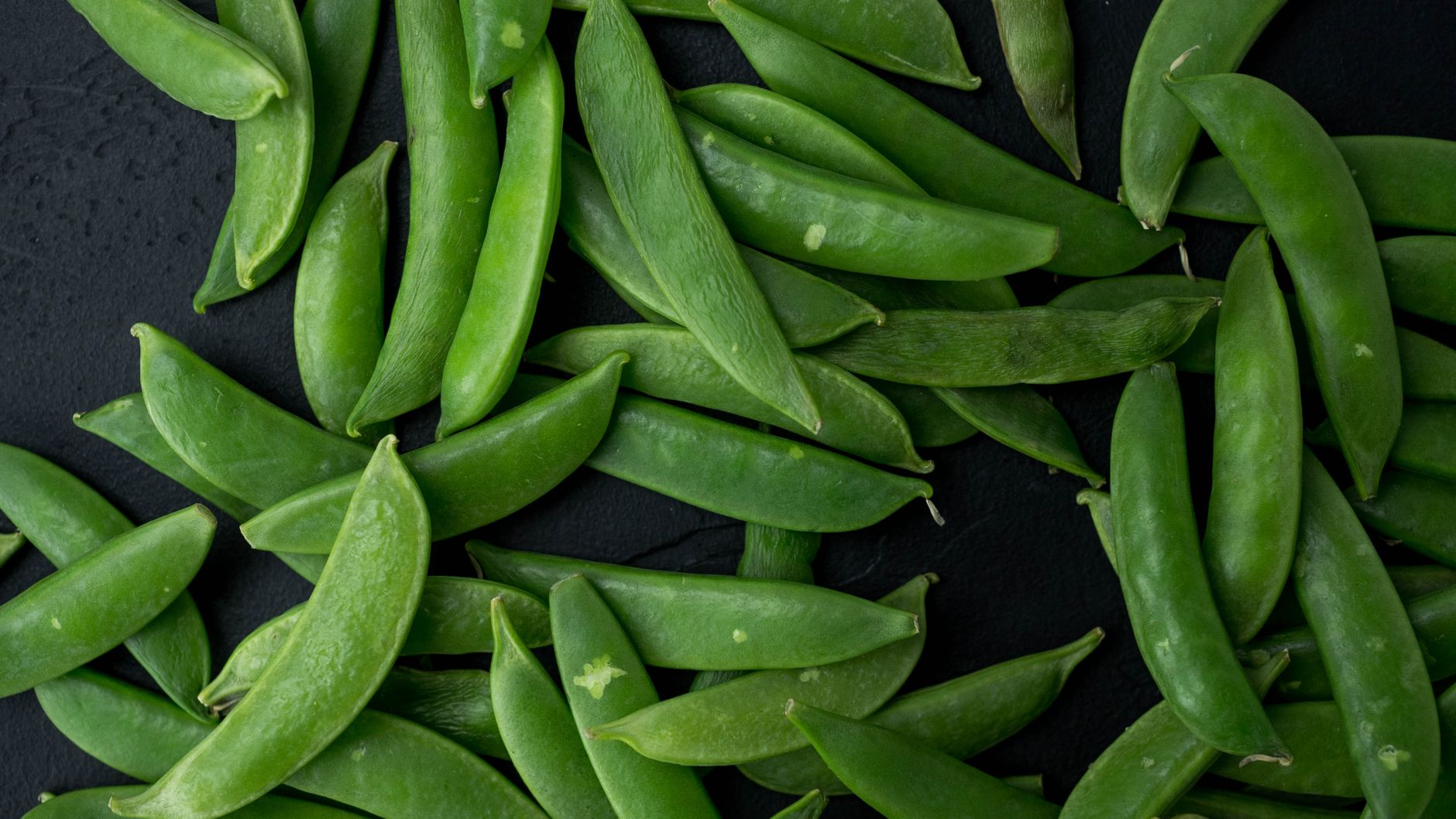 A close-up view of fresh green snap peas scattered on a dark surface, showcasing their vibrant color and texture.